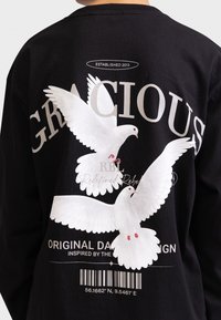 Fekete, hosszú ujjú póló, fehér galamb grafikával, a "KEDVES" felirattal és vonalkóddal; puha pamut anyag.