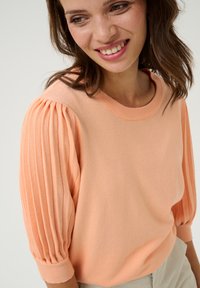 Pull en rose poudré avec un col rond côtelé, des manches bouffantes présentant des plis verticaux, et une texture en maille douce.