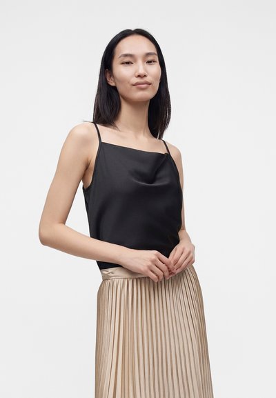 Femme asiatique portant un haut noir sans manches et une jupe plissée beige brillante, debout devant un fond clair uni.