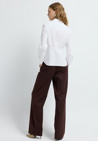 Donna con capelli mossi, che indossa una camicia bianca attillata con bottoni, pantaloni a righe marroni a gamba larga, tacchi beige, con le mani nelle tasche, rivolta di lato.