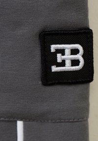 Grauer Stoff mit feiner Textur, versehen mit einem schwarzen gestickten Patch, der am Rand ein weißes "EB"-Logo zeigt. Saubere Nähdetails sind sichtbar.