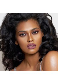 Close-up van een model met volumineus, donker krullend haar. Ze heeft een warme huidtint, gedurfde oogmake-up en een matte mauve lipkleur. Heldere huid.
