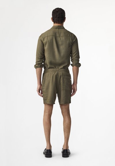 Homme debout de dos, portant une chemise olive à boutons avec manches remontées, un short assorti à taille élastique et des chaussures habillées noires.