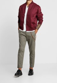 Burgundská bomber bunda s žebrovanými manžetami, bílá košile a světle zelené kalhoty. Černé boty s jemným leskem.