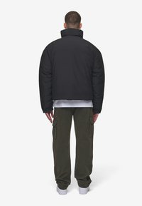 Sort polstret jakke med høj krave, båret over en hvid skjorte, parret med olivenfarvede cargo bukser og hvide sneakers. Bagvyn.
