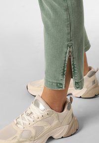 Gröna beskurna byxor med en dragkedja vid fållen, i kombination med ljusbeiga sneakers med nät- och läderdetaljer.