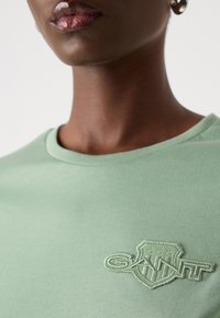 T-shirt in cotone verde chiaro con colletto rotondo e logo ricamato "GAINT" a forma di scudo sul lato sinistro. Texture liscia.