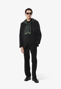Traje negro ajustado con una camisa negra, pantalones negros y zapatos negros. Bufanda verde atada al cuello, gafas de sol con lentes oscuros.