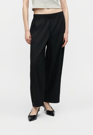 Γυναίκα που φοράει μαύρο παντελόνι με φαρδιά μπατζάκια και ελαστική μέση, μαζί με μαύρες γόβες μυτερές, στέκεται μπροστά σε λευκό φόντο.