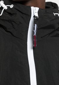 Czarna kurtka przeciwwiatrowa z białym zamkiem błyskawicznym i metką Tommy Jeans. Posiada lekką, pomarszczoną teksturę oraz regulowany kaptur z sznurkami.