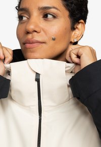 Lichtcrème jasje met zwarte mouwen en kraag. Heeft een reliëf "ROXY" logo, een zichtbare rits en een zachte, gladde textuur.