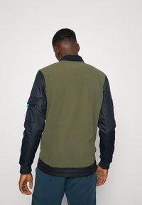 Zelená bomber bunda s tmavě modrými rukávy, žebrovanými manžetami a pasem. Na levém rukávu je boční kapsa s tyrkysovým akcentem.