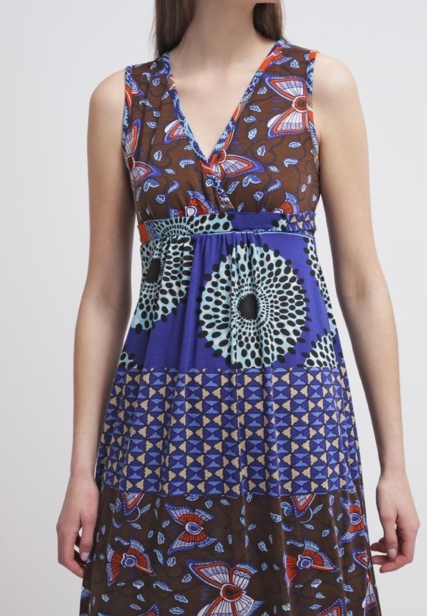 Femme portant une robe sans manches avec des motifs floraux et géométriques marron, bleu et orange, debout devant un fond clair uni.