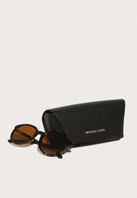 Skilpaddebrune solbriller med brune linser i et svart skinnetui merket "MICHAEL KORS". Etuiet har en avrundet design og en teksturert overflate.