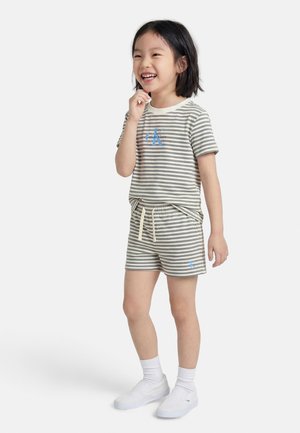 Smilende barn står og har en grå og hvidstribet Calvin Klein t-shirt og shorts på med hvide sokker og slip-on sko.