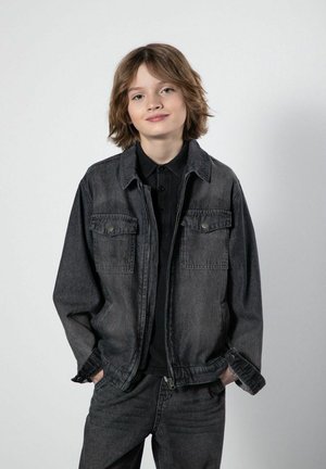 Młoda osoba z włosami do ramion, ubrana w czarną dżinsową kurtkę i spodnie, stojąca z rękami w kieszeniach na jednolitym tle.