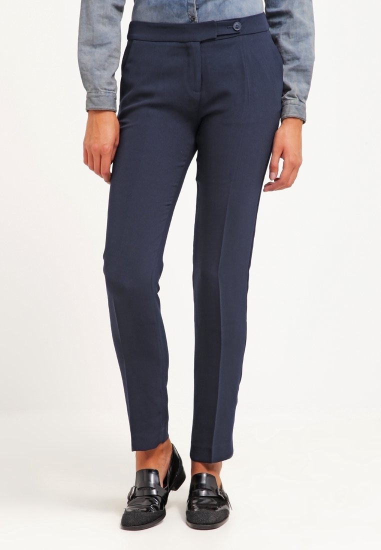 Personne portant un pantalon ajusté bleu marine foncé, une chemise bleue claire boutonnée et des mocassins noirs brillants, debout sur un fond blanc.
