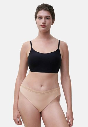 Frau trägt ein schwarzes, kurzes Camisole mit dünnen Trägern und hautfarbene Hipster-Unterwäsche mit Spitzenbund, steht vor einem einfachen Hintergrund.