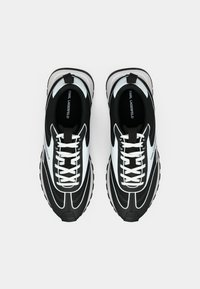 Crno-bijele atletske tenisice s teksturiranim materijalom, zakrivljenim dizajnom, istaknutim vezicama i debelim gumenim potplatom. Naziv marke na jeziku tenisice.