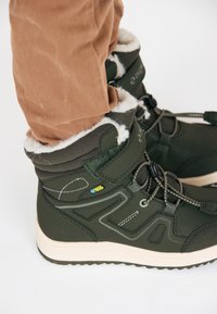 Dunkelgrüne High-Top-Stiefel mit weißem Fleece-Innenfutter, strukturiertem synthetischem Material, gepolstertem Schaft und kontrastierender heller Sohle.