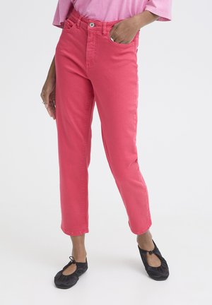 Personne portant un pantalon court rose, une chemise rose et des ballerines noires avec un nœud, debout avec une main dans la poche sur fond blanc.