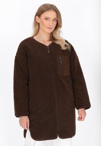 Cappotto in pile marrone con zip frontale, vestibilità rilassata, maniche lunghe e taschino sul petto. Texture morbida con una finitura superficie shaggy.