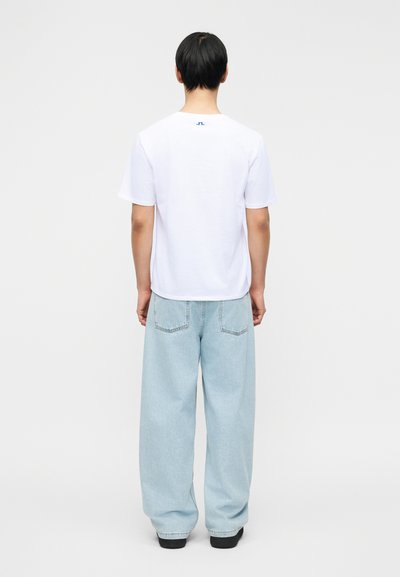 Άνδρας με κόμη προς τα πίσω φοράει άσπρο κοντομάνικο πουκάμισο με μικρό μπλε λογότυπο και ανοιχτόχρωμο μπλε άνετο τζιν με μαύρα παπούτσια.