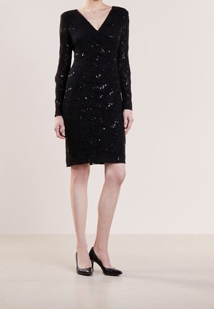 Femme portant une robe noire en dentelle longueur genou avec des manches longues et des sequins subtils, associée à des chaussures noires à talons hauts.