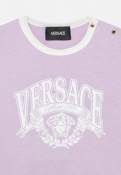 T-shirt lilas Versace avec col blanc, deux clous dorés sur l’épaule et grand logo blanc avec tête de Méduse et texte « Established 1978 ».