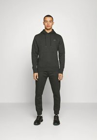 Pelēks kokvilnas džemperis ar kanguro kabatu un aukliņām, kopā ar tumši pelēkām sporta biksēm. Apģērbu papildina melni sporta apavi.