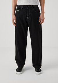Schwarze Jeans im lockeren Schnitt, mit weißen Nähten und einem grünen Etikett an der vorderen Tasche. Kombiniert mit schwarzen Sneakers.