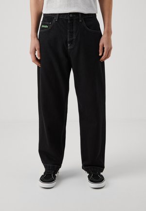 Black denim jeans with a loose fit, white stitching, and a green label on the front pocket. Paired with black sneakers.