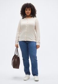 Light beige cardigan with button closure, long sleeves, and soft texture, paired with blue jeans and a brown woven tote bag. White sneakers complete the look.