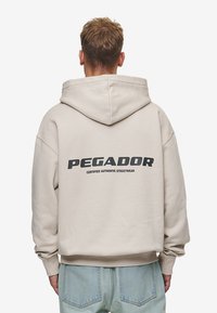 Bež majica s kapuljačom s velikim crnim logom "PEGADOR" na leđima, s kapuljačom na vezanje i rebrastim manžetama, u kombinaciji sa svijetlim trapericama.