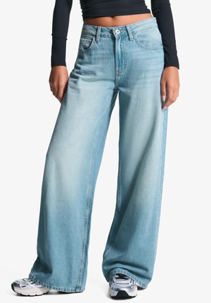 Femme portant un jean bleu clair à jambes larges, un crop top noir à manches longues et des baskets argentées, debout devant un fond blanc.