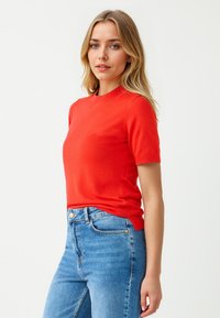 Rode gebreide top met korte mouwen en een ronde halslijn, gemaakt van een gladde stof. Draagt bij blauwe high-waisted jeans met een sluiting aan de voorkant.