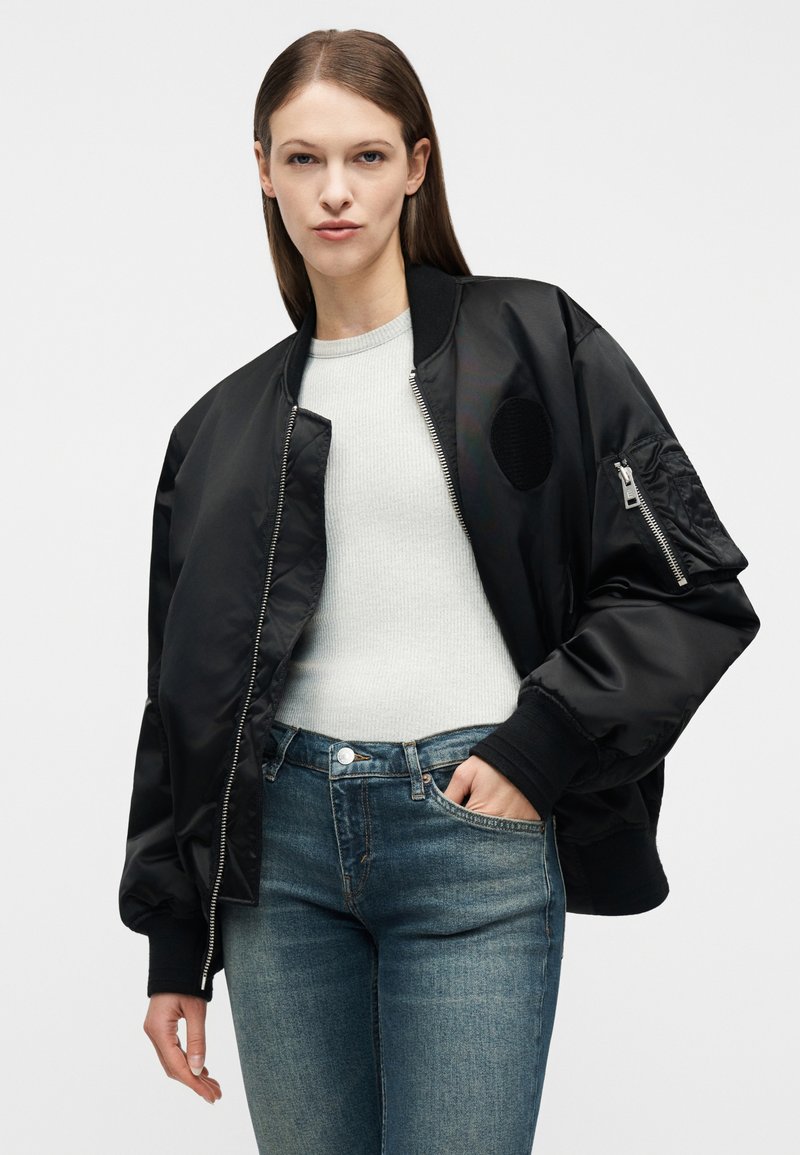 Frau trägt schwarze Bomberjacke mit Reißverschluss und einem Aufnäher am Ärmel über weißem, geripptem Oberteil und blauen Jeans, Hand in der Tasche, neutraler Hintergrund.