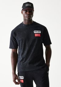 Μαύρο κοντομάνικο μπλουζάκι με υφή, με λευκά και κόκκινα λογότυπα. Συνδυάζεται με μαύρη φόρμα και μαύρο καπέλο.