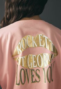 Rožinės spalvos medvilniniai marškinėliai su auksinio metalo spalvos spauda lūpų formos. Tekste yra užrašas „ROCKETT ST GEORGE LOVES YOU“ įvairių šrifto dydžių.