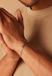 Silberne Kettenarmband an einem Handgelenk, bestehend aus miteinander verbundenen Gliedern mit polierter Oberfläche. Die Hände sind vor einem neutralen Hintergrund zusammengelegt.