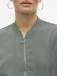 Gros plan sur une femme portant des boucles d'oreilles créoles en or et une veste bomber vert sauge avec une fermeture éclair argentée et un col côtelé.