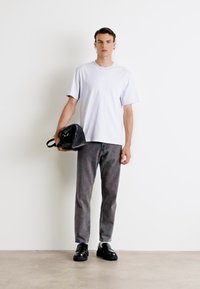 T-shirt blanche à manches courtes, pantalon gris, chaussures noires. Le modèle tient un sac noir, debout devant un fond blanc uni.
