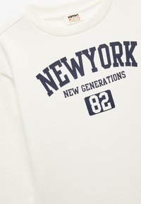 Felpa bianca con stampa blu navy. Presenta le scritte "NEW YORK," "NEW GENERATIONS," e "82." Collo a costine, tessuto morbido, vestibilità ampia.
