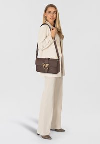 Borsa a spalla in pelle marrone con hardware oro, forma rettangolare, chiusura a scatto e finitura testurizzata, indossata con un completo crema.