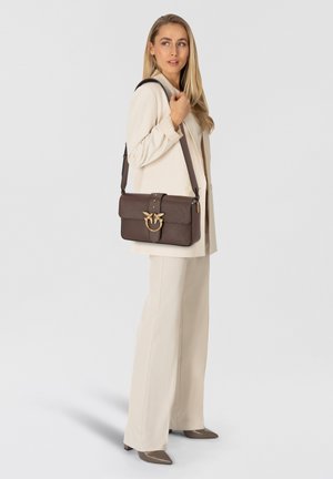 Borsa a spalla in pelle marrone con hardware oro, forma rettangolare, chiusura a scatto e finitura testurizzata, indossata con un completo crema.