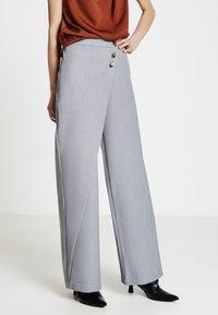 Pantaloni grigi chiari a gamba larga con una texture liscia, doppio bottone frontale e vita alta, abbinati a tacchi neri con punta a mandorla.