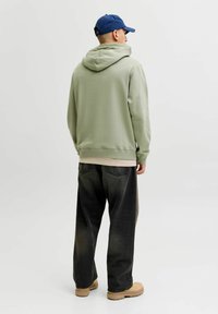 Vaaleanvihreä huppari rentoa istuvuutta, yhdistetty tummiin leveälahkeisiin farkkuihin ja vaaleisiin kenkiin, vaaleaa valkoista taustaa vasten.