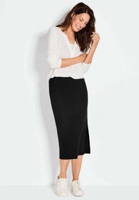 Jupe noire longueur genou en tissu extensible avec une fente sur le côté, portée avec un haut blanc à manches longues. Associée à des baskets blanches.