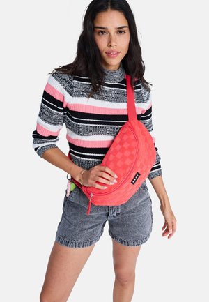 Frau trägt einen schwarz-weiß-pinken gestreiften Pullover mit grauen Jeansshorts und hält eine rote, strukturierte Umhängetasche quer über der Brust.