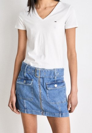 Wit v-hals T-shirt gecombineerd met een blauwe denim minirok met een rits aan de voorkant, twee voorzakken en een riem met een zilveren gesp.